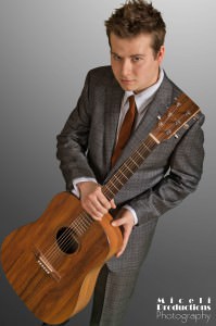 Photo of Musician Aaron Fry, in a gray suit standing with his guitar