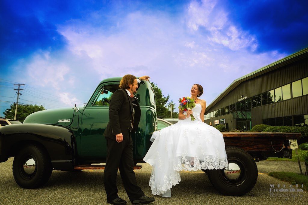 Bride and groom couple share a moment sitting on an old Chevy pick-up truck with a bright blue sky on their wedding day.