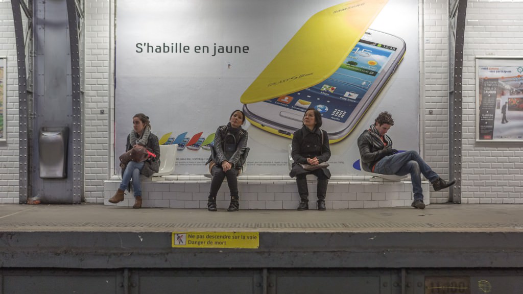 Four individuals sit in a Paris train station waiting for the subway train. They are all evenly spaced, doing their own activities.