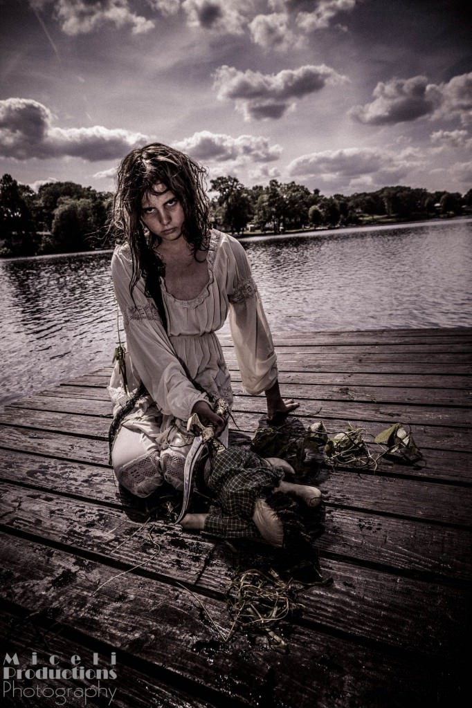 Halloween photography shoot. Scary image of a young girl that crawled out of a lake and is holding a knife over a voodoo doll.
