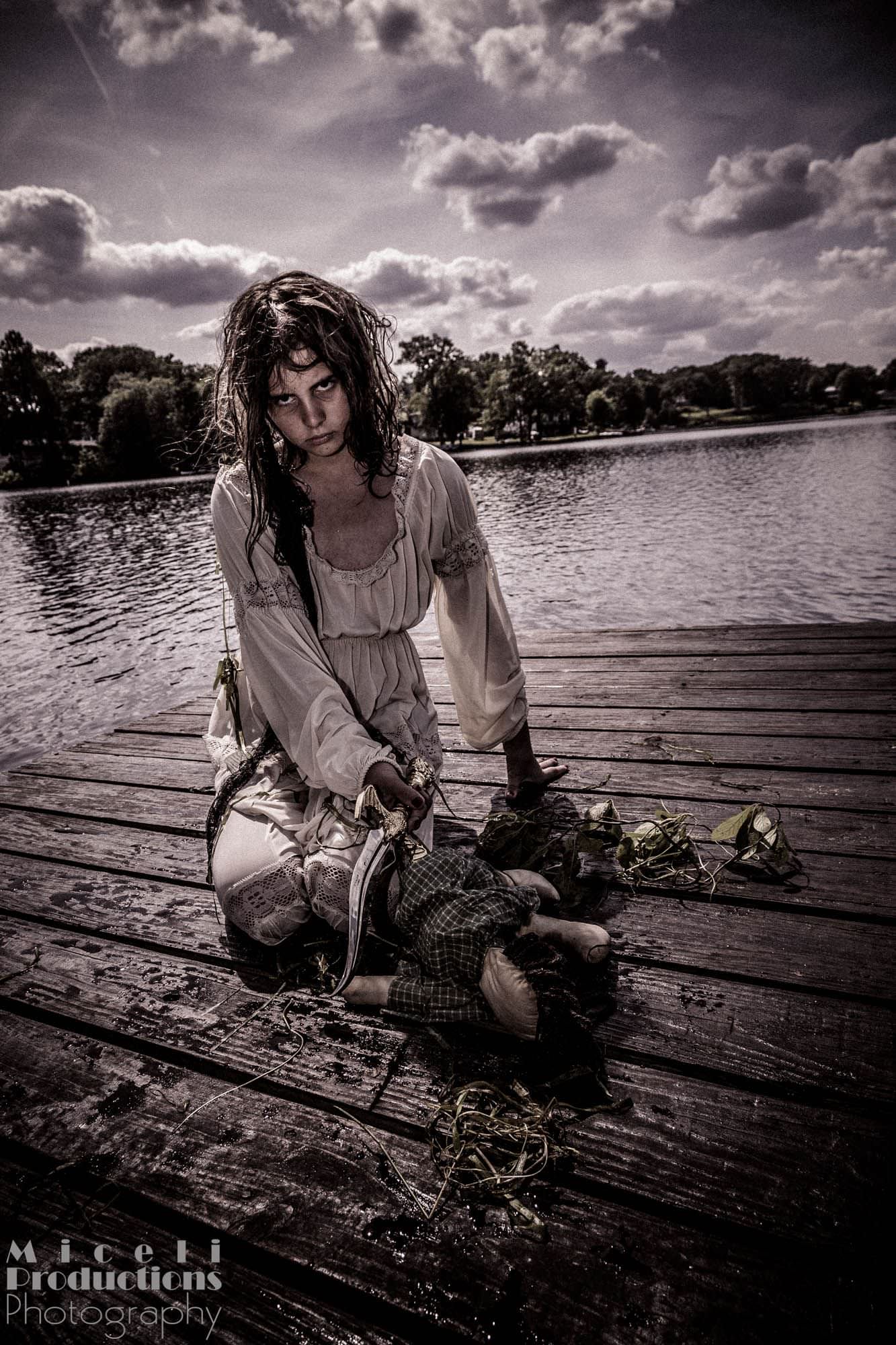 Halloween photography shoot. Scary image of a young girl that crawled out of a lake and is holding a knife over a voodoo doll. Photo by Miceli Productions