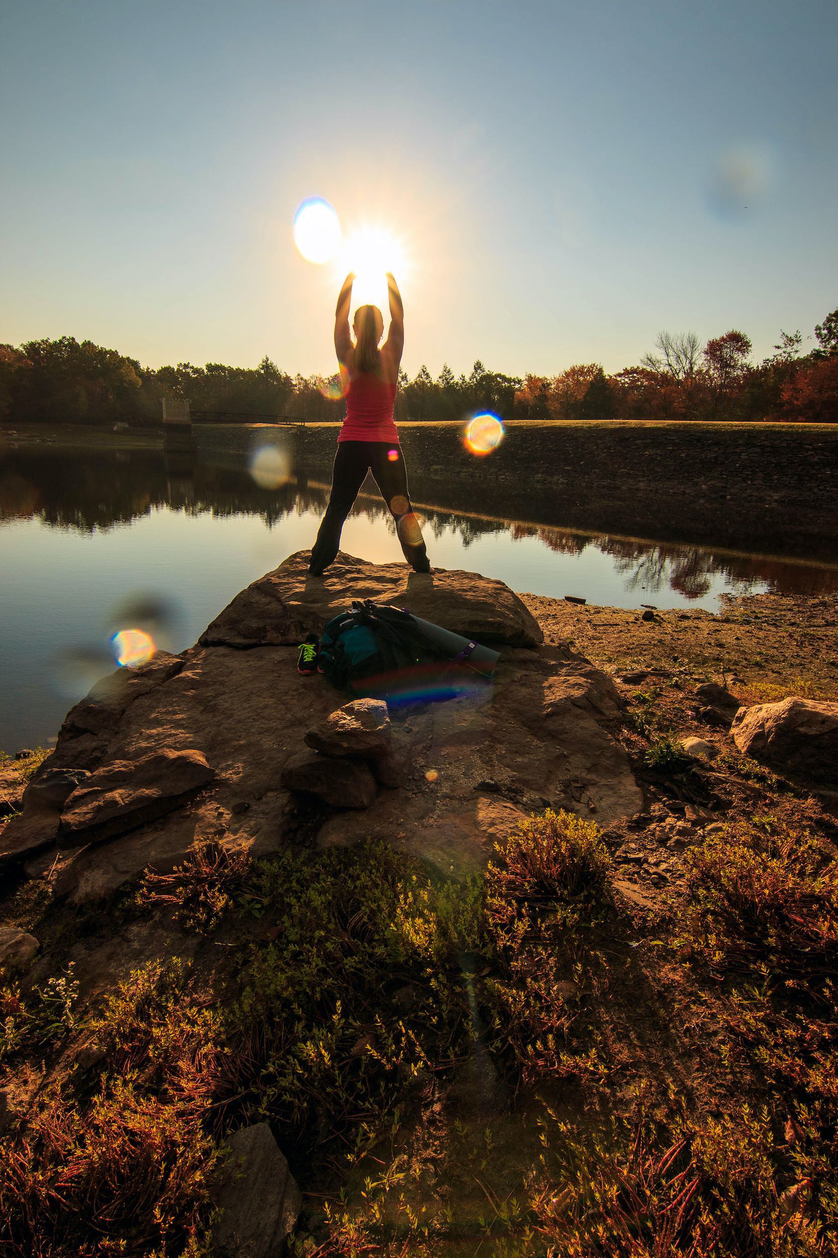 Photo of early morning sunrise with girl holding the sun in her up stretched hands while standing on a rock outcropping near a lake.
