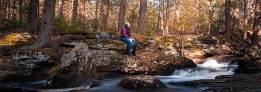 Woman hiker sits on a rock overlooking an evergreen forest stream, Mad River, in Connecticut.